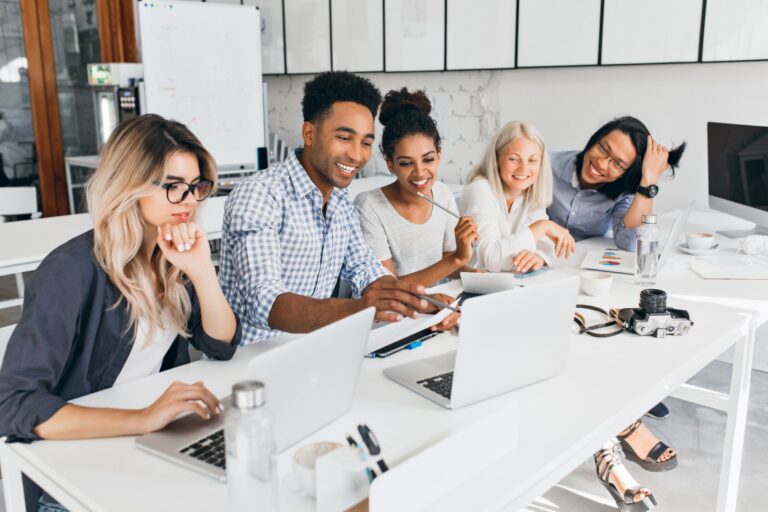 Smiling african student pointing with pencil at laptop screen. Concentrated blonde girl in glasses propping chin with hand while working with computer in office..